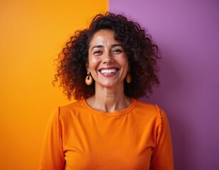 Fototapeta premium Middle aged hispanic woman with curly hair smiles broadly. She wears an orange shirt against a vibrant orange and purple background. Her expression is joyful and confident.