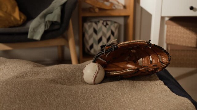 No people close-up of brown leather baseball glove and ball lying on bed in teenage boys bedroom, armchair and drawer in background, suggesting sport hobby, training or physical activity