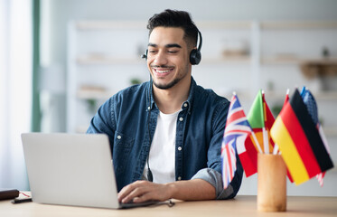 Happy arab guy student learning foreign language online, sitting at workdesk and using laptop, headset, typing on keyboard and smiling. Middle eastern man teacher having video chat with student © Prostock-studio
