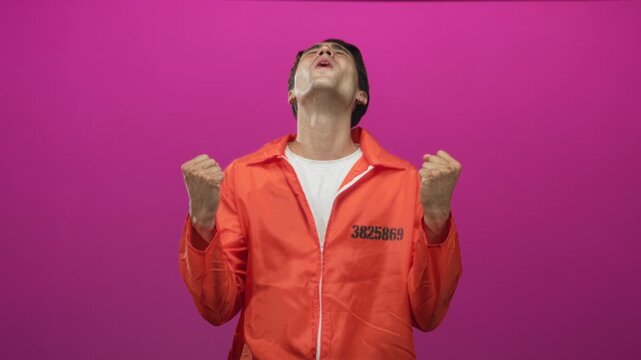 Man prisoner wearing orange jumpsuit with visible inmate number clenches fists, tilts head back and mouths a shout in studio; relief redemption.