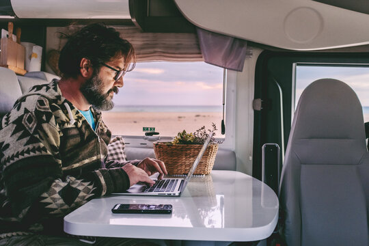Adult man typing on laptop inside a motorhome parked on a sandy beach, remote work with ocean view, embracing van life freedom and a mobile office lifestyle