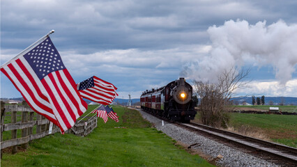 A steam train moves along railroad tracks in a countryside setting. American flags are planted along the route, and there are clouds overhead. The scene is lively with movement and nature. © Greg Kelton