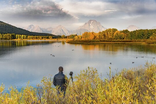 Photographer standing with tripod at the river, Snake River, Mount Moran at the back, morning mood at Oxbow Bend, autumn trees, Grand Teton Range, Grand Teton National Park, Wyoming, USA