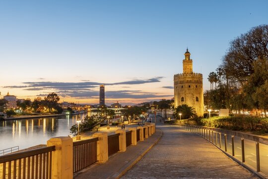 Boardwalk at the river Rio Guadalquivir with illuminated Torre del Oro, in the back Torre Sevilla, sunset, blue hour, Sevilla, Andalusia, Spain