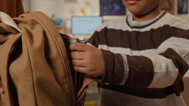 Medium close-up of 6-year-old African American boy packing schoolbag with textbooks, copybooks while getting ready to start elementary school, putting smartphone in pocket, supported by older sibling