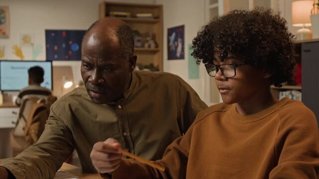 Medium close-up of attentive black father explaining teenage son grammar rules using online educational recourse on laptop, while writing test or homework, boy understanding and exclaiming with joy