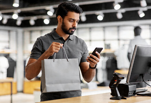 Indian Man Holding Grey Shopping Bag Looking at Smartphone at Retail Store Checkout Counter Modern Indoor Lighting