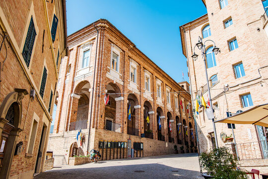 Corinaldo historic town hall building with arcades and flags, standing majestically on an ancient cobblestone street in a charming Italian village against a clear blue sky