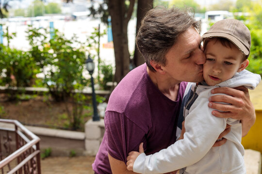 father kissing little son on cheek while hugging him outdoors in city park, happy family moment and parental love, dad and child concept