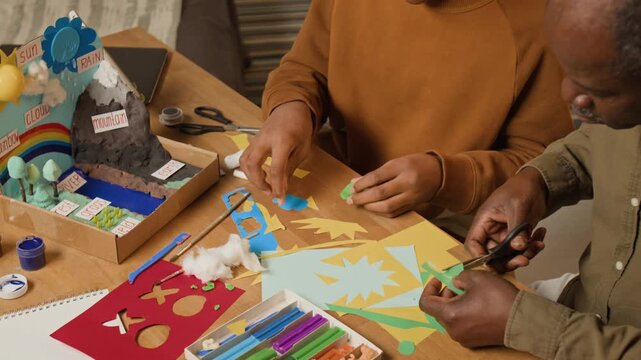 High-angle arc shot of African American father and son creating dimensional project for science lesson at school, representing Earth, atmosphere, climate, environment from color paper and clay