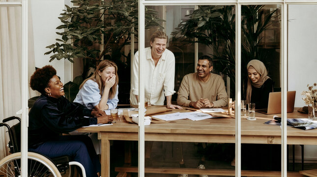 Joyful diverse group of five architects laughing and discussing plans around long wooden table in glass atrium with wheelchair user, embodying inclusive, happy teamwork in modern office.

