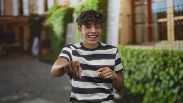 Teenage man pointing finger at camera, laughing and winking in a striped tshirt by a hedge and building facade on a sunlit street courtyard; playful.