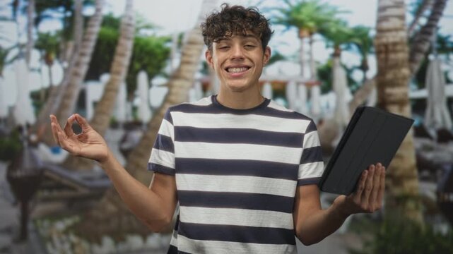 Teenage boy holding tablet and making ok sign beside palm trees and pool at resort building; relaxation mindfulness.