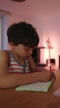 Young boy sitting at table doing homework in evening light, slightly frustrated expression while working through exercise, quiet moment of effort and concentration at home