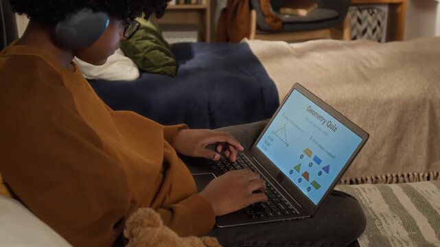 Medium side shot of focused teenage African American boy in headphones sitting on bed at home with laptop, performing online geometry test while studying on remote edtech e-learning platform