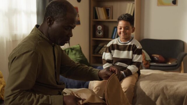 Medium shot of caring African American dad packing schoolbag for little son in bedroom in morning, putting in lunchbox, urging not to be late for lesson, boy shaking head, refusing to go to school