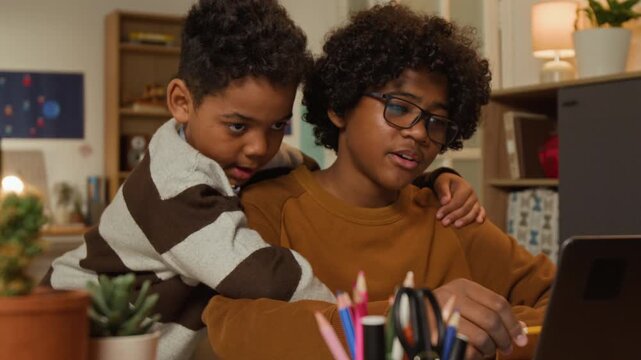 Medium shot of 13-year-old African American schoolboy sitting at desk with laptop, working on study or coding project, explaining to curious younger brother watching while hugging him on shoulder
