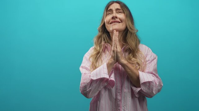 Woman with hands clasped in prayer looking upward against turquoise studio backdrop; hopeful earnest plea.