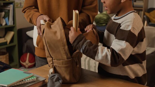 Medium close-up of African American boy assisting young brother with preparing for beginning elementary school, gathering textbooks and pencil case into backpack and zipping