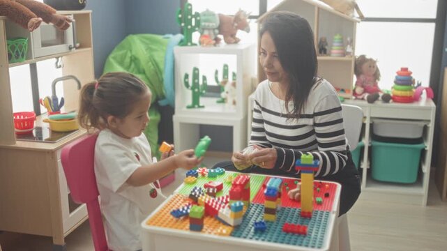 Woman handing and placing toy bricks, stacking towers with little girl at colorful play table in studio; learning bonding warmth.