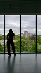 Fototapeta premium Young woman stands by a large panoramic window in a modern office looking at the city view. Vertical full length silhouette shot. Concept of future planning, career goals and urban lifestyle