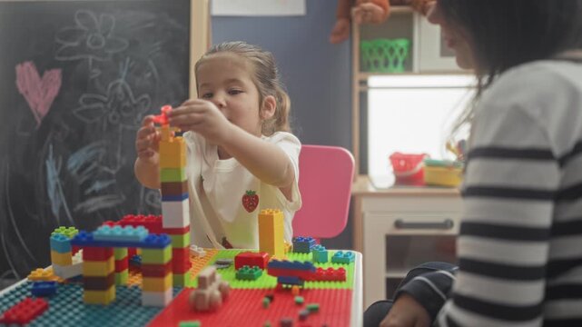 Girl stacking colorful building blocks on a tall tower at a table in classroom, placing bricks while adult guides nearby; concentration learning.