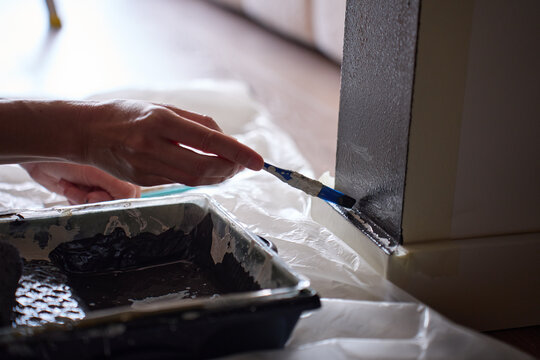 Hand applies dark paint with small brush along wall edge beside tray and plastic sheet