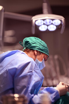 Female Surgeon Performing Surgery In Operating Room Under Surgical Light Wearing Scrubs And Mask