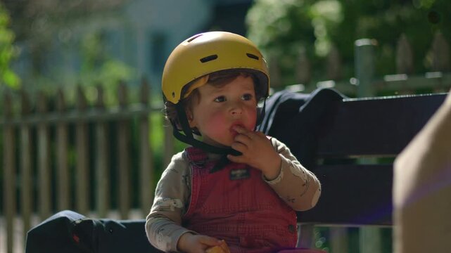 Toddler in yellow helmet seated on park bench, nibbling tangerine slices in soft sunlight, candid moment of quiet focus and sensory discovery during outdoor play