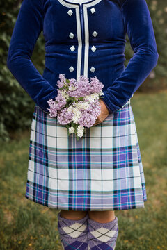 Highland dancer in blue velvet jacket and purple tartan kilt holding bouquet of spring lilacs