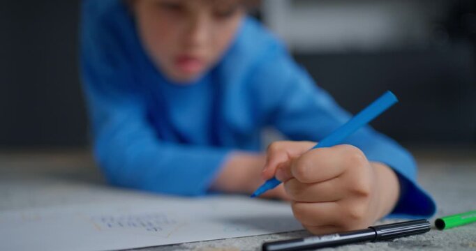 Focused young child in blue shirt writing on paper with colorful pens during quiet homework or drawing practice at home, close-up study scene