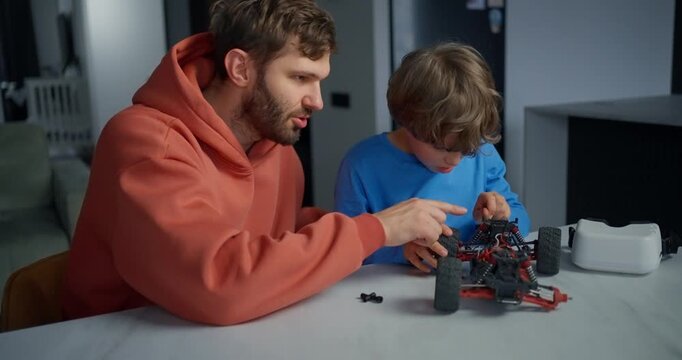 Father and young son assembling a remote control toy car together at a kitchen table, sharing a focused hands-on learning moment indoors
