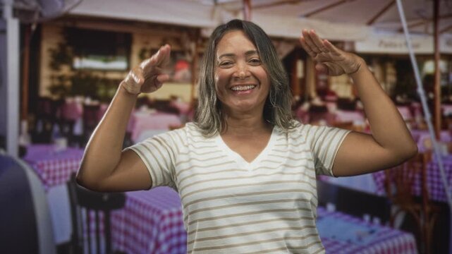 Woman frames face with hands and smiles while seated at an outdoor cafe on street with checkered tables and chairs; joy playfulness.
