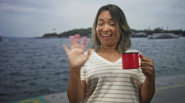 Hispanic woman waving hand while holding red cup on a seaside dock with boats and ferry visible in the distance; cheerful greeting.