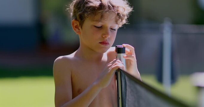 Shirtless boy examining swimming pool safety fence post while standing outside in backyard during warm sunny day with focused expression