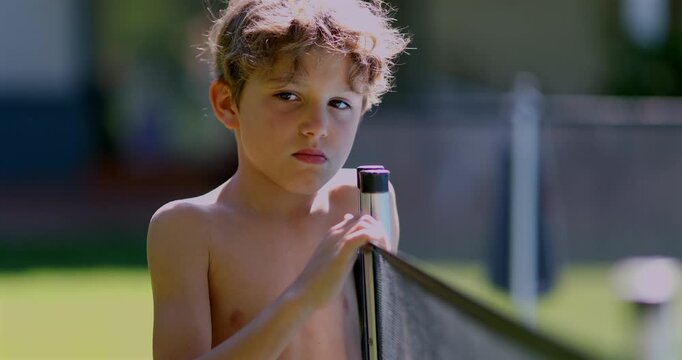 Shirtless boy standing beside swimming pool safety fence with thoughtful expression, hands holding post on sunny day in backyard outdoor area