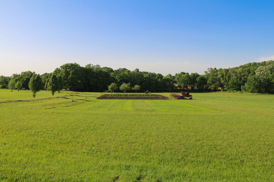 A green field stretches beneath a clear blue sky with a small garden patch trees in the distance and red farm equipment on a sunny day in the countryside