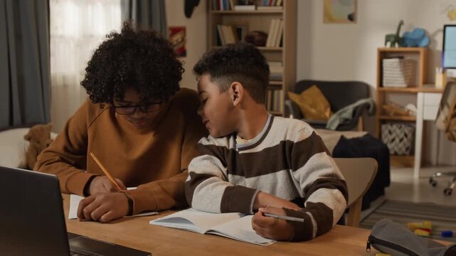 Medium shot of cheerful African American teenage and elementary school student brothers doing homework together, sitting side by side at desk, peeking at each others copybooks, pointing at mistakes