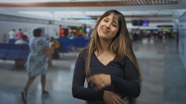 Young woman with hand to chin and crossed arm in an airport terminal, thoughtful gaze upward and subtle smile; contemplation.