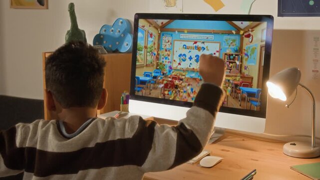Medium rear shot of young African American boy cheering and whooping with joy after giving correct answer while doing math sums on online app on computer in bedroom, preparing for elementary school