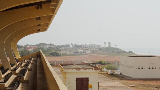 Abandoned Cape Coast Sports Stadium in Ghana with empty stands revealing coastal city skyline and Atlantic Ocean on hazy day in West Africa showing poverty
