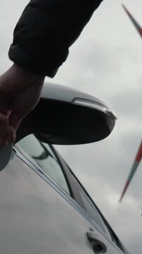 Vertical video of male hands disconnecting wind power plug of charging station from electric car. Unrecognizable man unplugs charging cable from electric car near generator windmill turbine