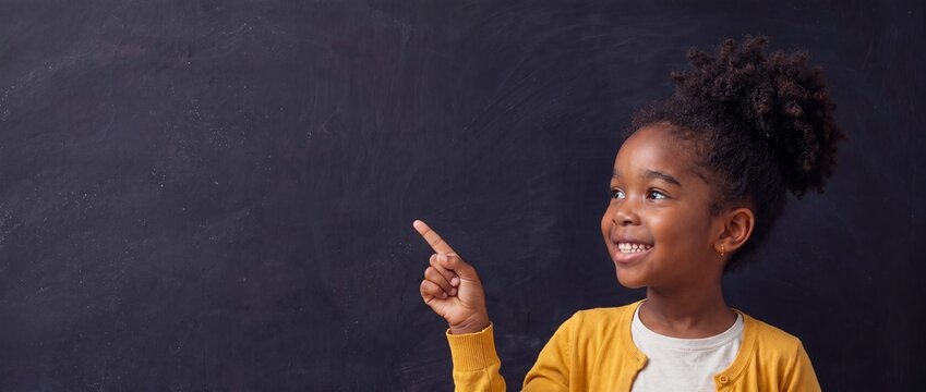 Back to school concept: Smiling African American black schoolgirl pointing finger aside at an empty blackboard. Long Banner with copy space for text.