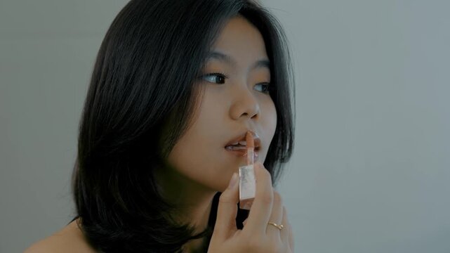 Side view of pretty young asian woman applying lip gloss make up cosmetic preparation. Happy beautiful female model looking the mirror apply lips balm to getting ready in the morning, beauty routine.