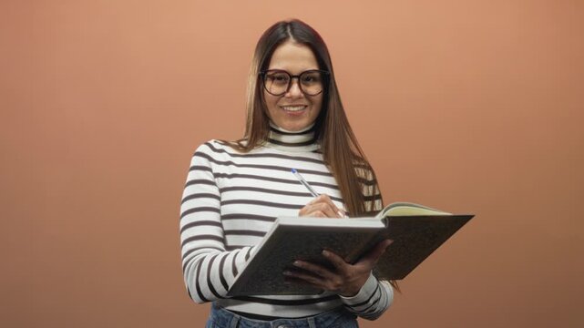 Young hispanic woman writing in a notebook, holding pen and open book, wearing glasses and striped turtleneck in studio; focused study.