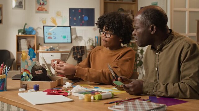 Medium shot of mature African American father and teenage son crafting creative handmade natural science project together for school, cutting up color paper with scissors and discussing ideas