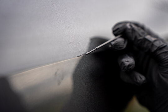 Hand in a black nitrile glove using a microbrush to apply touch up paint to a deep scratch on a car body, performing professional automotive paint chip and scratch repair.