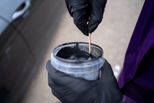 Hands in black nitrile gloves stirring silver metallic automotive paint in a plastic jar with a wooden stick, preparing car paint for refinishing or body repair service.
