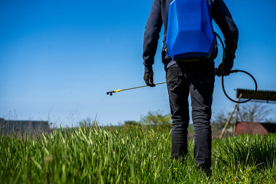 Worker with backpack sprayer applying herbicide or tick control chemical on wild green grass field during sunny daytime outdoors.