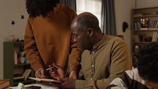 Medium shot of mature dedicated black father reviewing mathematics homework with teenage son, explaining mistakes, then turning to younger child learning to write, committed to education achievements